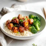 Crispy sesame tofu cubes glazed in a glossy sesame sauce served with rice and broccoli in a white ceramic bowl.